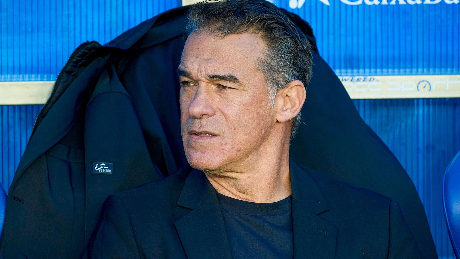 Luis García Plaza, con chaqueta negra y camiseta oscura, posa sentado en un banco. Detrás de él, un abrigo negro cuelga, con una pared azul como fondo.