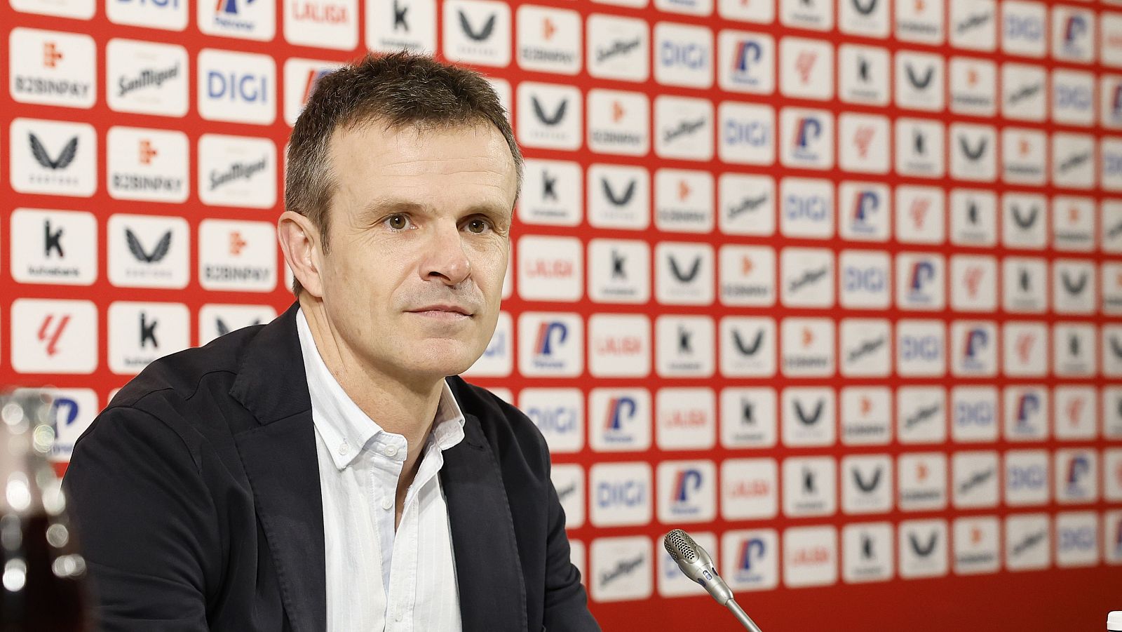 Jon Uriarte, presidente del club, habla en una rueda de prensa, con un micrófono y un panel de patrocinadores con logos como DIGI y San Miguel de fondo.