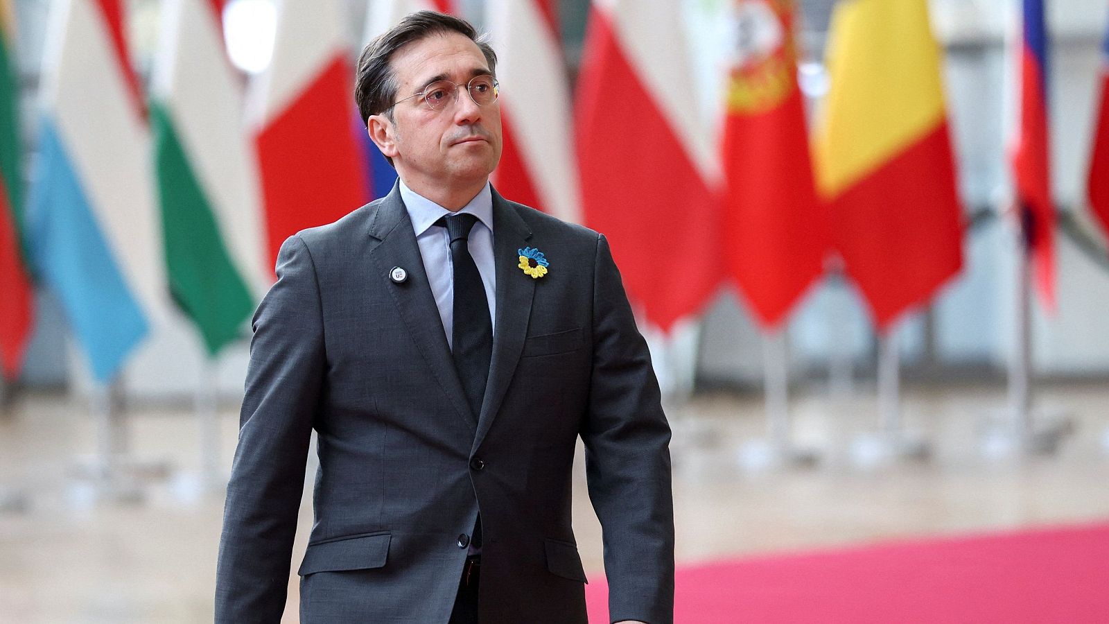 José Manuel Albares, con traje oscuro y gafas, posa frente a un conjunto de banderas de diversos países. En su solapa, se aprecian pines, uno con la bandera de Ucrania.