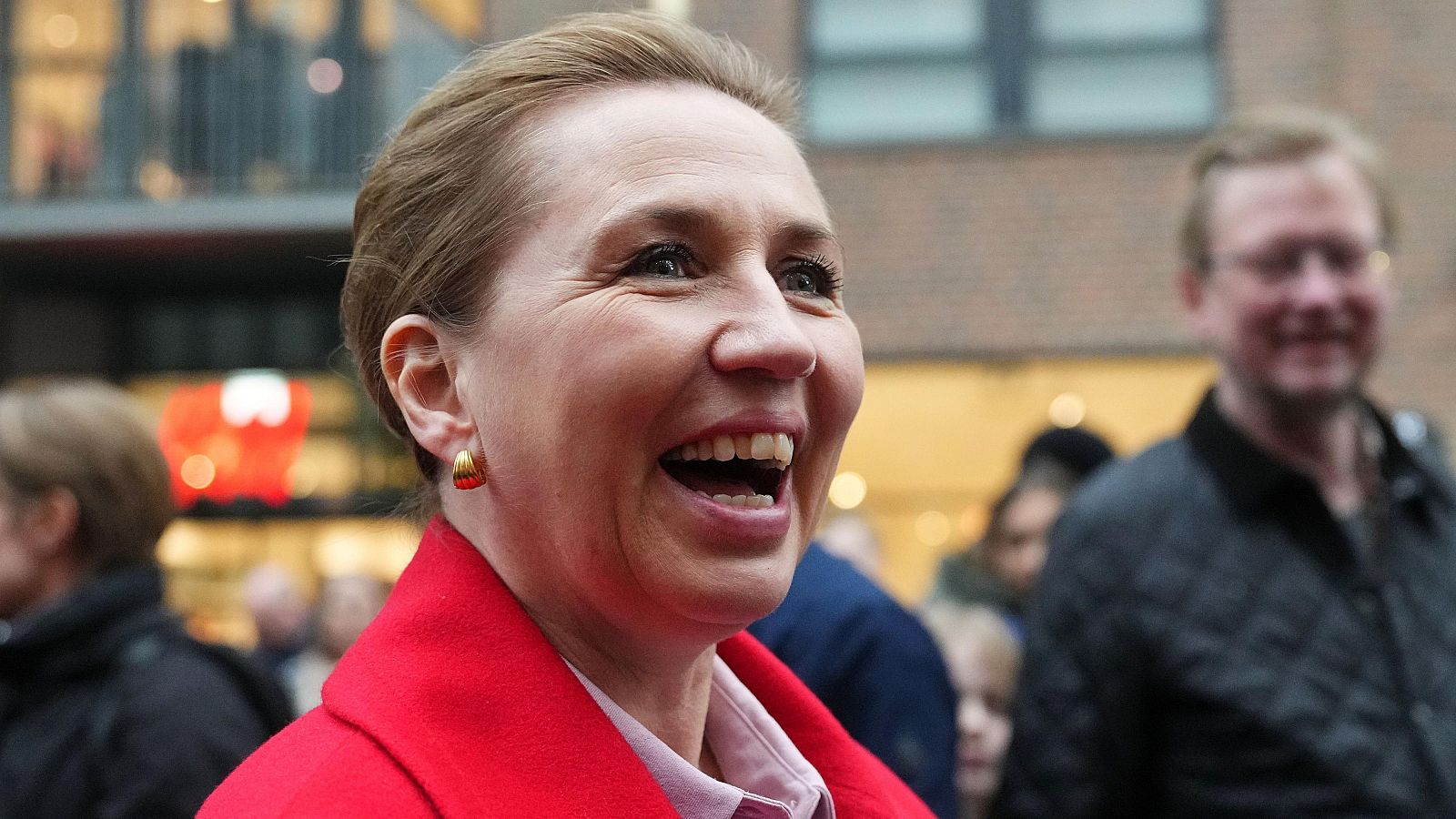 Mette Frederiksen, con abrigo rojo y pendientes dorados, sonríe en un entorno urbano. Se observa a personas desenfocadas detrás de ella.