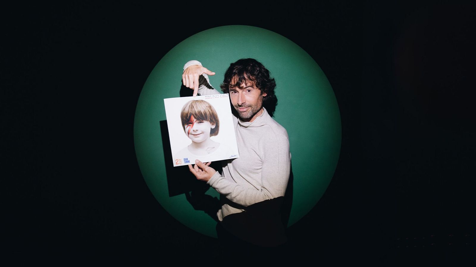 Bruno Oro, amb barba i jersei clar, mostra una portada de disc amb la imatge d'un nen amb un llamp vermell a la cara, en un fons negre amb un cercle verd.