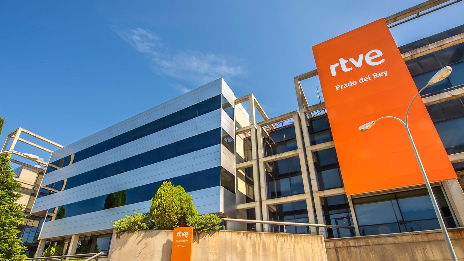 Vista exterior de los edificios de RTVE en Prado del Rey, con dos estructuras principales: una moderna y otra con una estructura más abierta. Un letrero naranja con el logo de RTVE y la inscripción "Prado del Rey" destaca en la fachada.