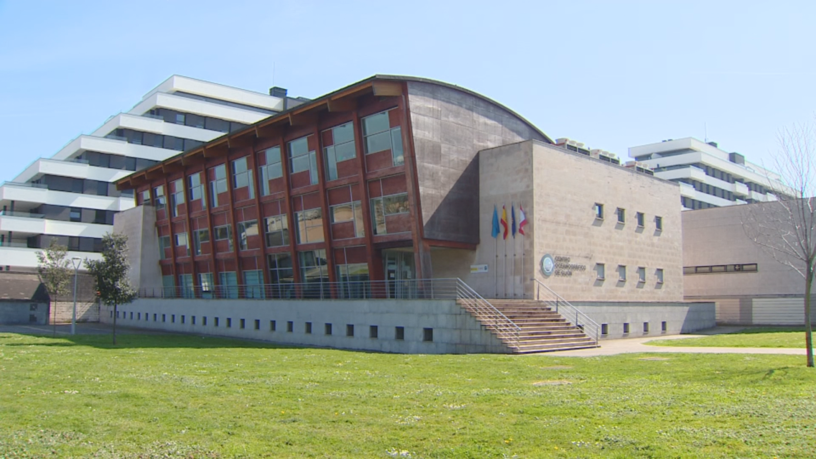 El edificio principal del centro, con una arquitectura moderna, presenta una sección de madera con ventanales y otra de hormigón con banderas y un logotipo, precedida por una escalera.