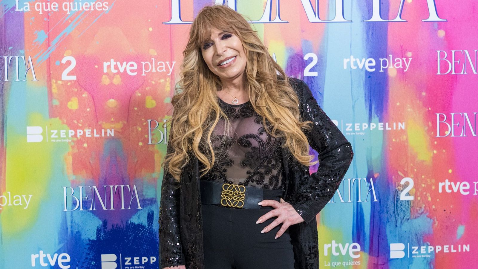 Una mujer con cabello rubio y ondulado posa frente a un fondo colorido con textos promocionales. Viste una blusa negra, chaqueta brillante, pantalones negros y un cinturón con un logo dorado.