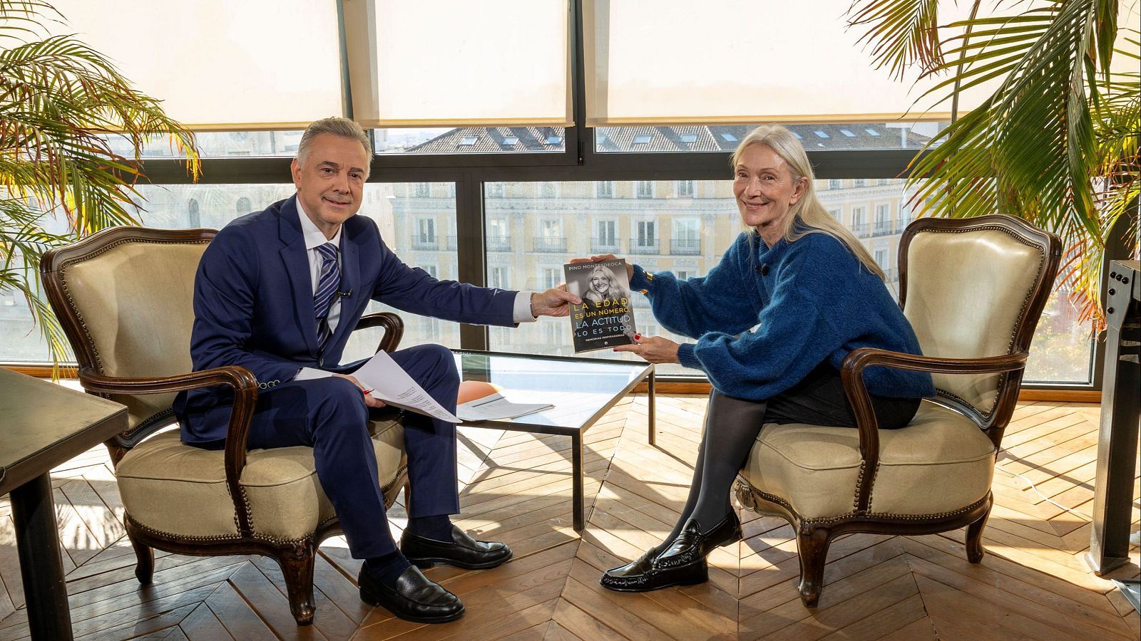 Jenaro Castro, con traje azul, entrega un libro a Pino Montesdeoca, quien viste un jersey azul, en un ambiente interior con ventanas y mobiliario clásico. El libro tiene una portada con una fotografía y un título.