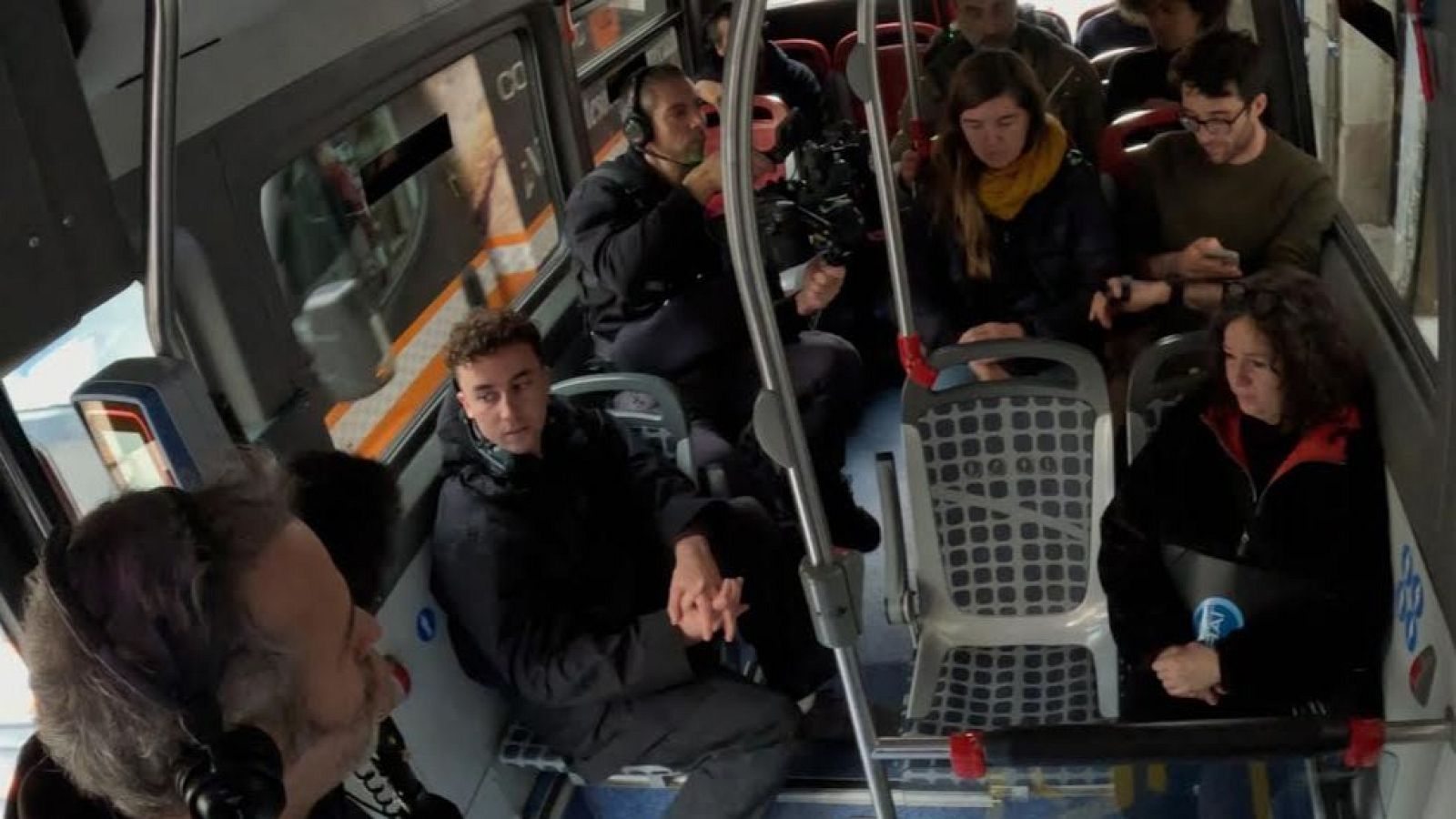 Dins d'un autobús, un home amb auriculars i càmera grava mentre altres passatgers, homes i dones, ocupen els seients i l'espai dempeus.