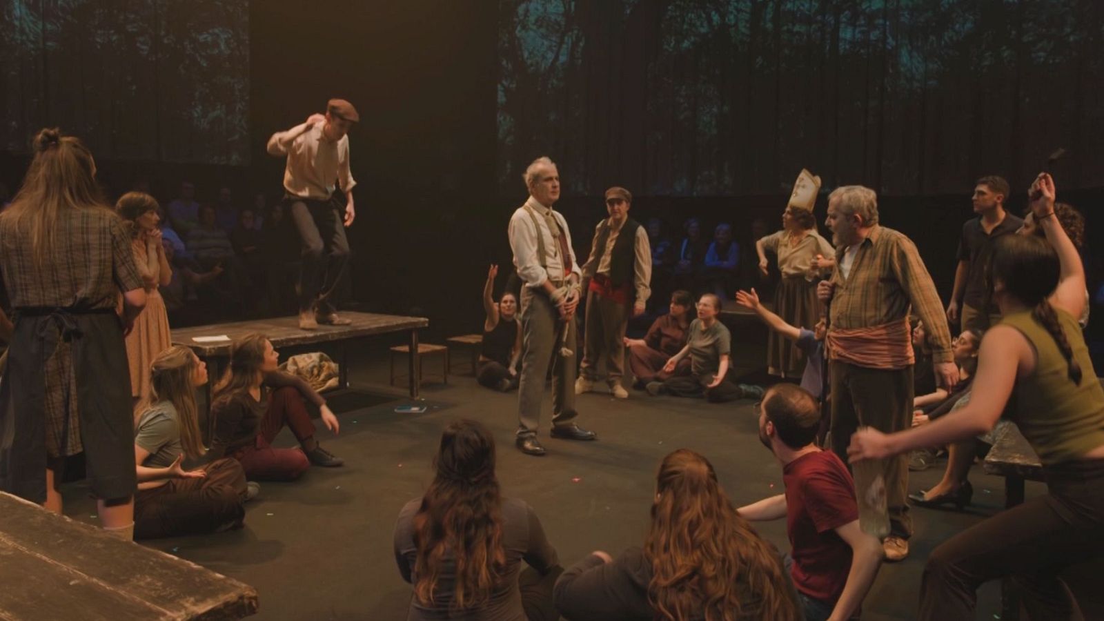 En un escenario iluminado con luz tenue, un joven actúa sobre una plataforma de madera, rodeado por un grupo de personas. Un hombre mayor con tirantes y otro con chaleco y gorra observan la escena, posiblemente dirigiendo la representación teatral.