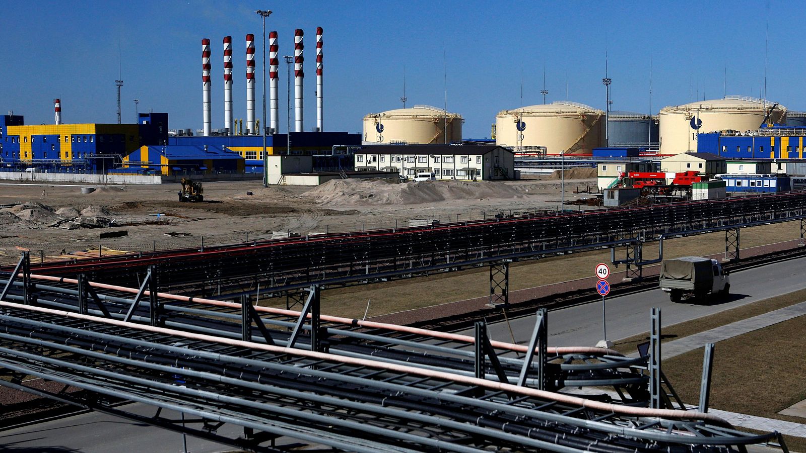 Una terminal industrial muestra tuberías, tanques de almacenamiento, edificios y una carretera con un camión, con chimeneas al fondo.