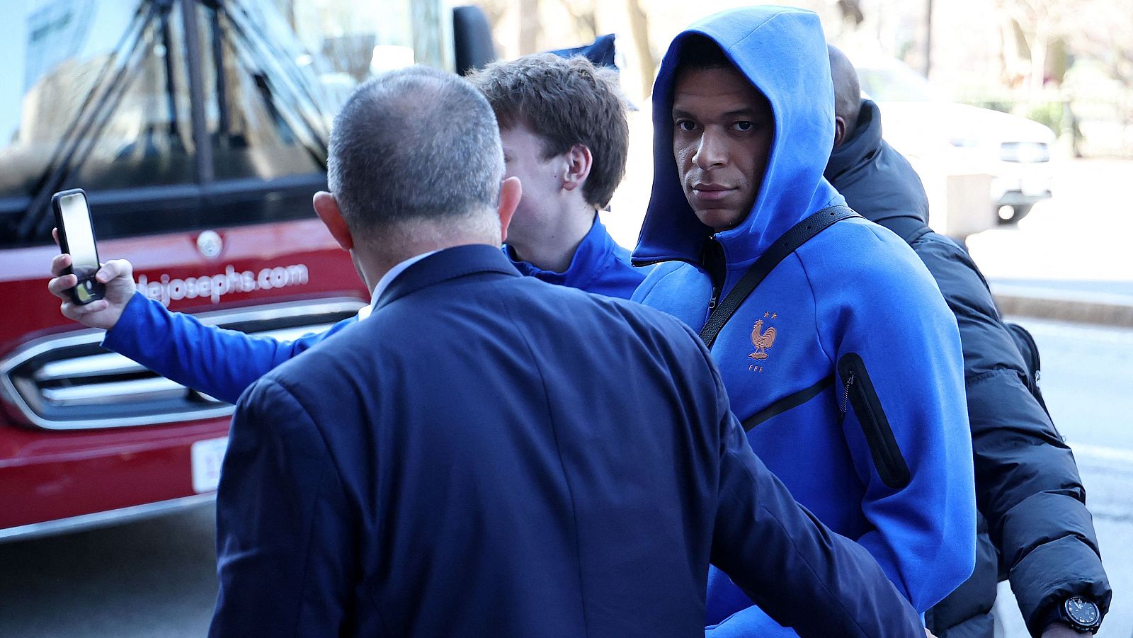 En la imagen, un futbolista con una sudadera azul es fotografiado por un hombre de traje. Un autobús rojo con una inscripción se encuentra en el fondo de la escena.