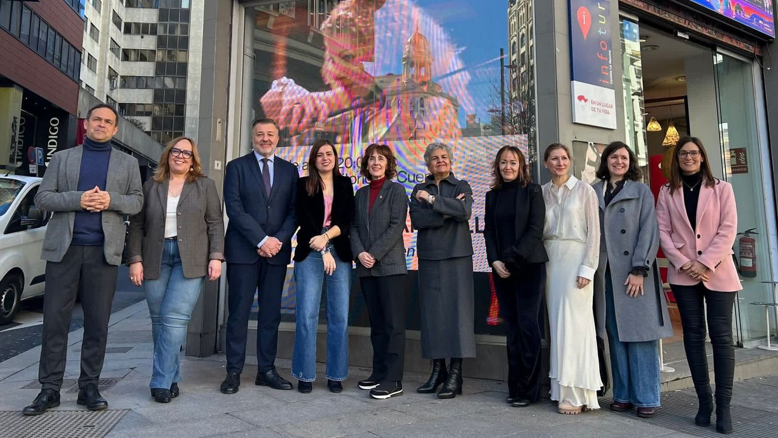 Un grupo de personas posa frente a un edificio, con una variedad de vestimentas que incluyen chaquetas, blusas y pantalones. Se observa una pantalla en el edificio y un letrero con información turística.