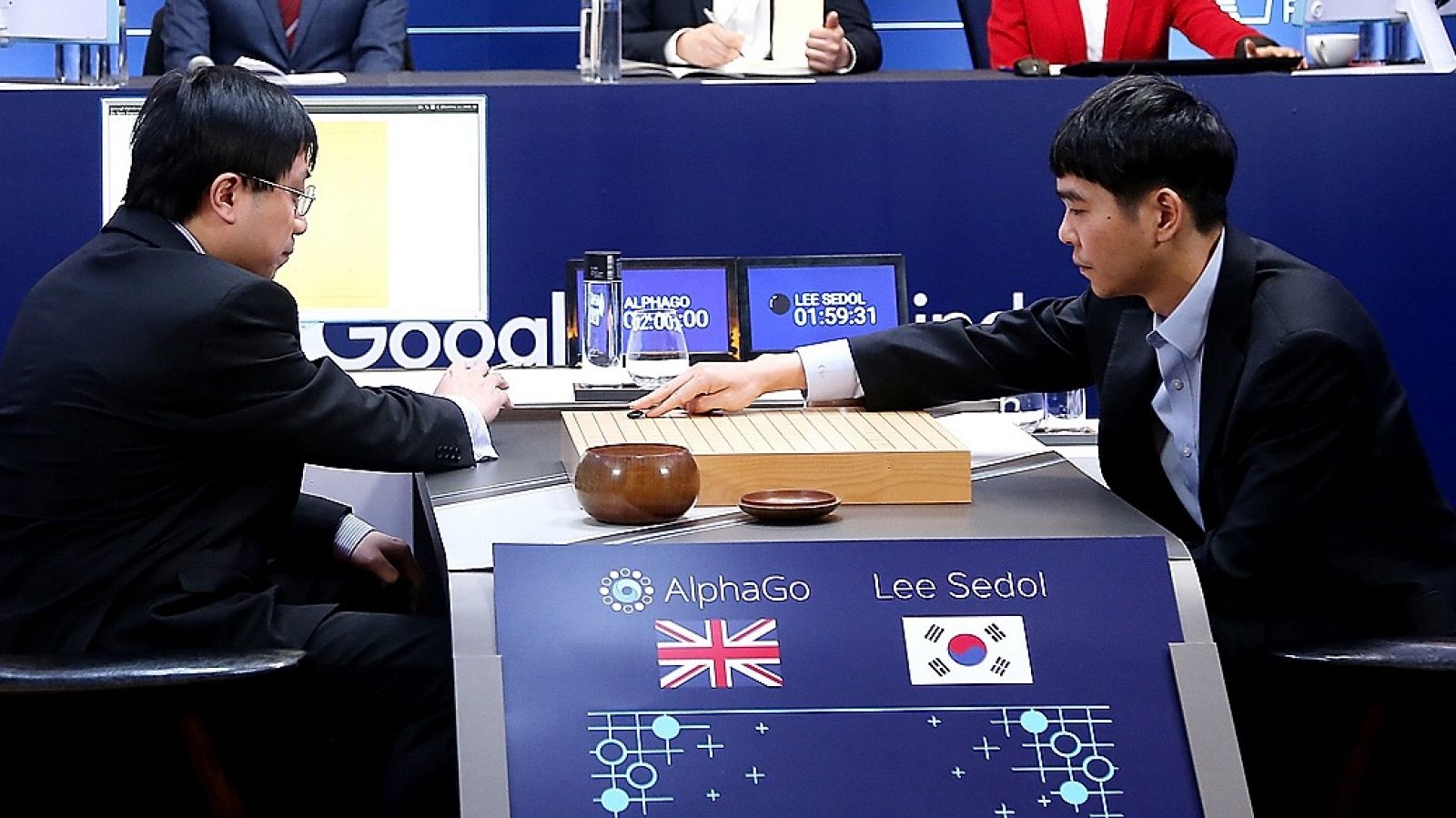 Un jugador realiza un movimiento en un tablero de Go frente a su oponente; en primer plano, un cartel con banderas identifica a “AlphaGo” y “Lee Sedol”.
