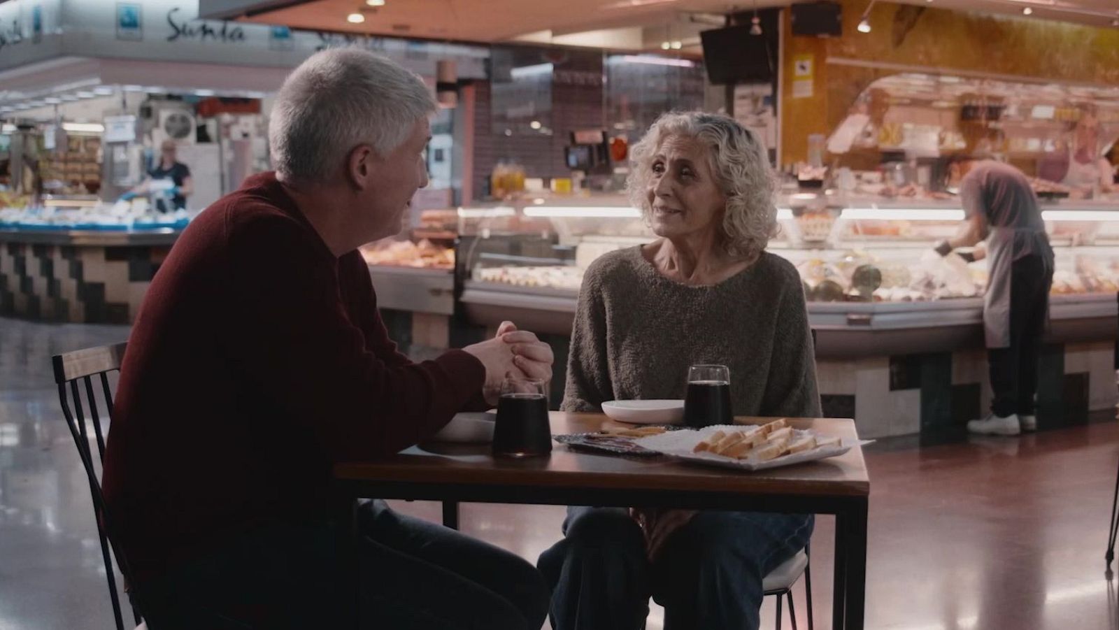Dues persones conversen assegudes a una taula en un mercat, amb begudes i menjar. Una d'elles, amb cabells arrissats, mira a l'altre, que té cabells canosos. Al fons, es veuen els taulells del mercat amb productes.