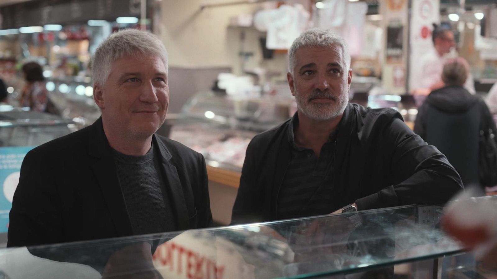 Dos homes amb cabells blancs, Pep Prieto i Adrià Collado, es troben al mercat. Un d'ells somriu i vesteix una jaqueta negra, mentre que l'altre porta un rellotge.