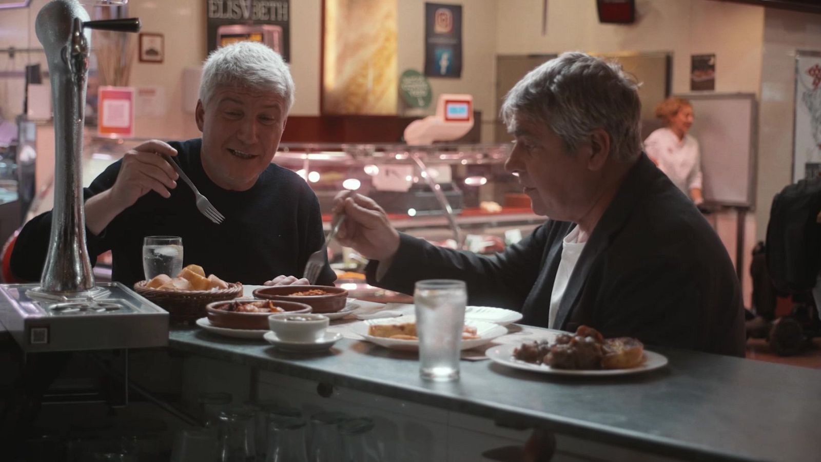 Dos homes, un amb samarreta negra i l'altre amb jaqueta fosca, mengen tapes i beuen aigua en un bar. A la barra hi ha diversos plats amb menjar i un cistell de patates fregides.