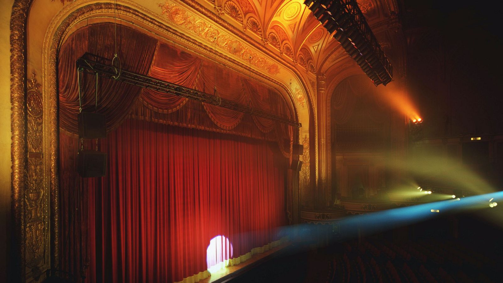 En el interior de un teatro, una cortina roja es el centro de atención, iluminada por un foco que crea un círculo de luz. La arquitectura clásica se complementa con detalles dorados y luces de colores que iluminan el espacio.