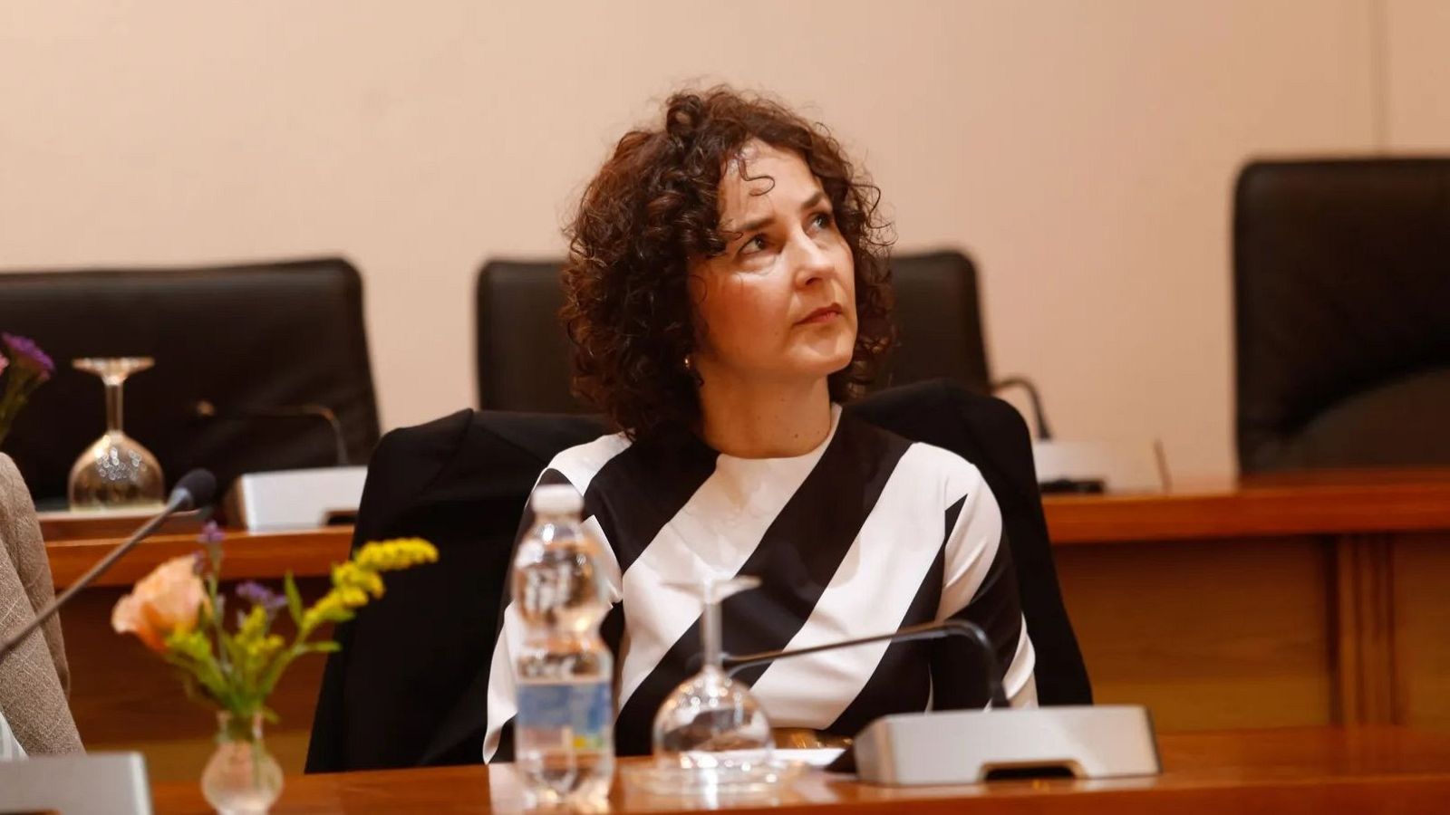 Mª Ángeles Martínez-García, subdirectora de la Cátedra RTVE-US, posa sentada en una mesa con un micrófono y una botella de agua. Lleva una blusa a rayas y el fondo muestra sillas.