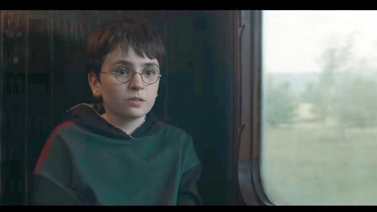 Dominic McLaughlin, caracterizado como Harry Potter, mira por la ventana de un tren. Lleva una sudadera verde con capucha y gafas redondas, con el pelo castaño oscuro.