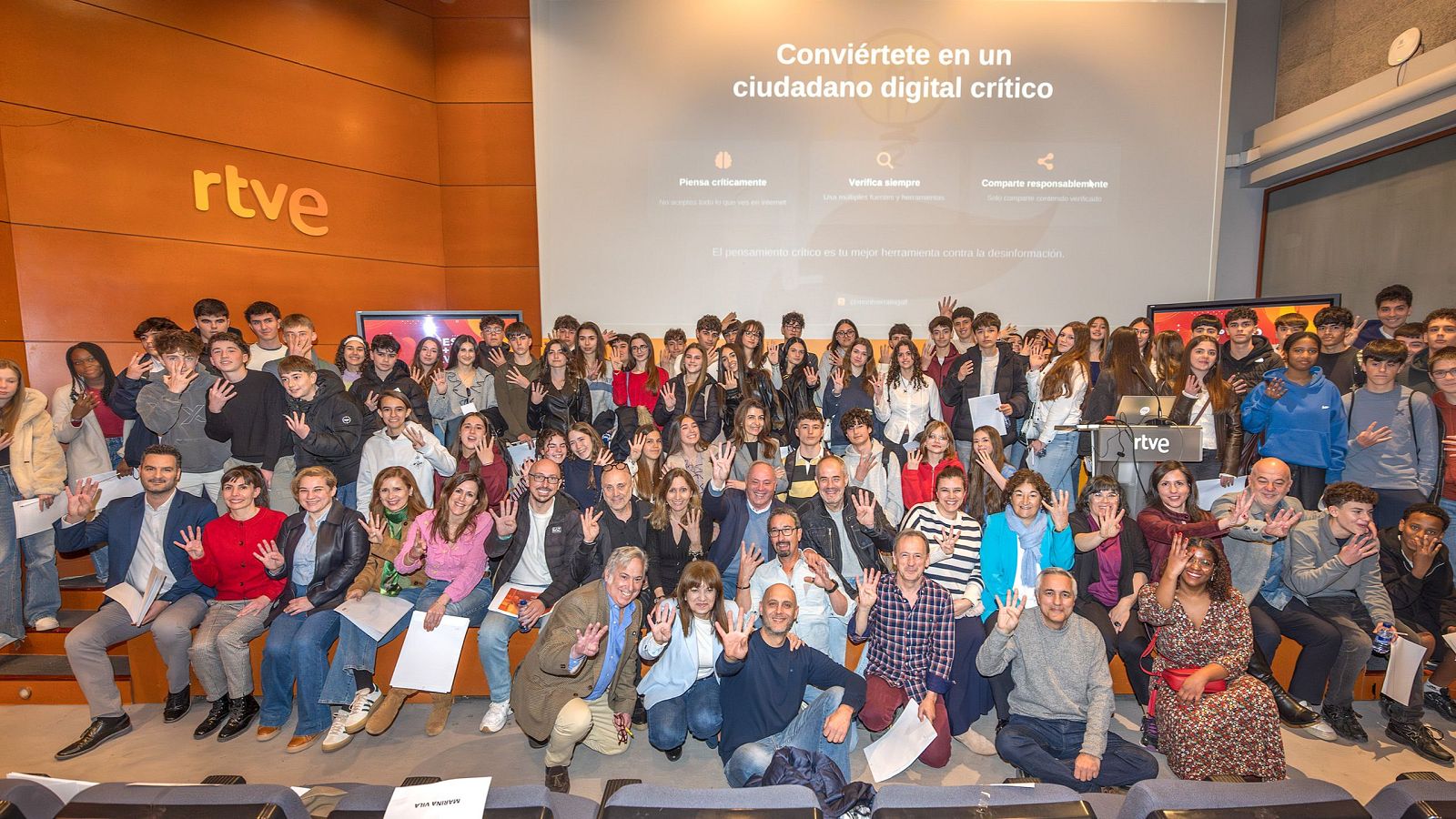 En un auditorio, un grupo de estudiantes de secundaria y adultos posan para una foto. Los jóvenes levantan la mano, mientras que al fondo se observa una pantalla con mensajes relacionados con el pensamiento crítico y el uso responsable de la información.