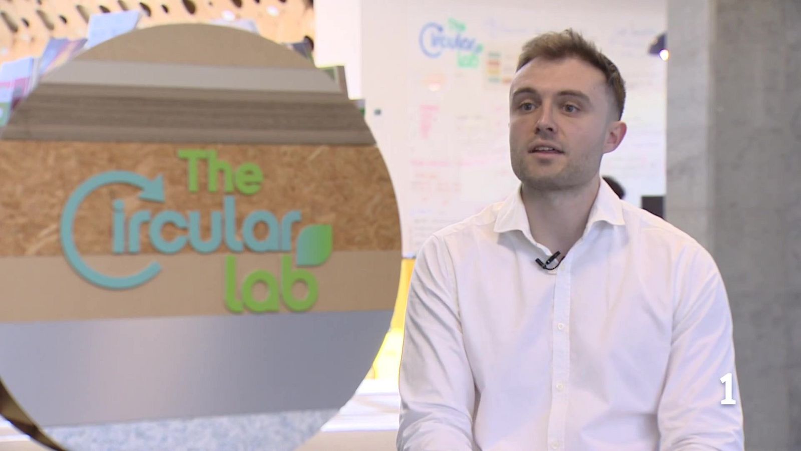 Un hombre joven con camisa blanca posa frente a una pared con texto y gráficos. A su izquierda, se observa un logo circular.
