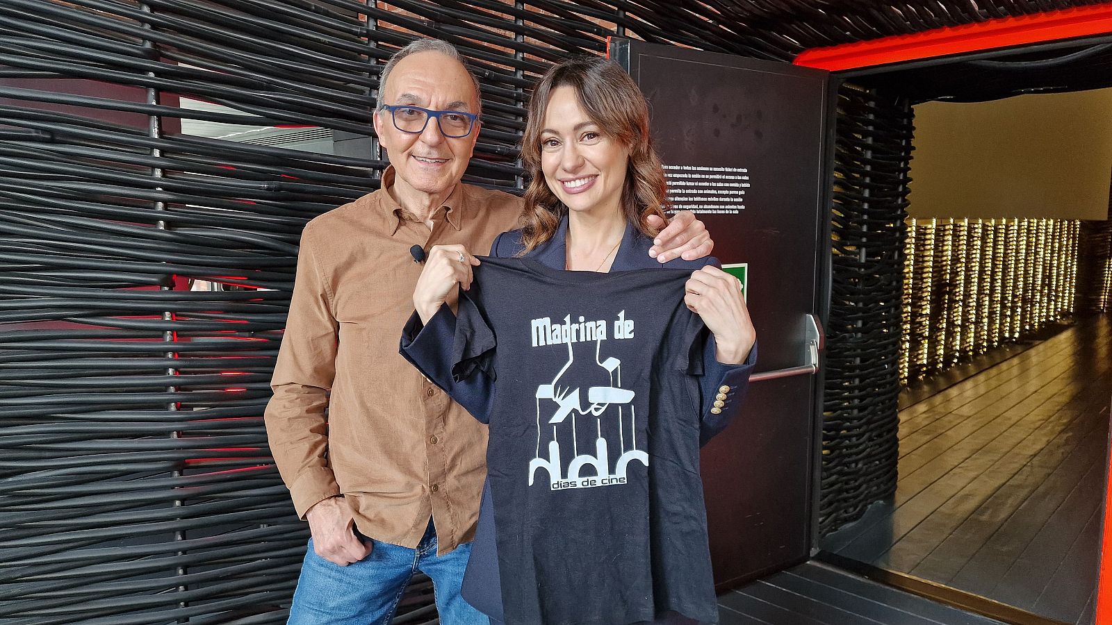 Natalia Verbeke, con una camiseta promocional, posa junto a un hombre. El hombre, con gafas, apoya su mano sobre el hombro de Natalia. El fondo muestra una pared con líneas horizontales y una puerta.