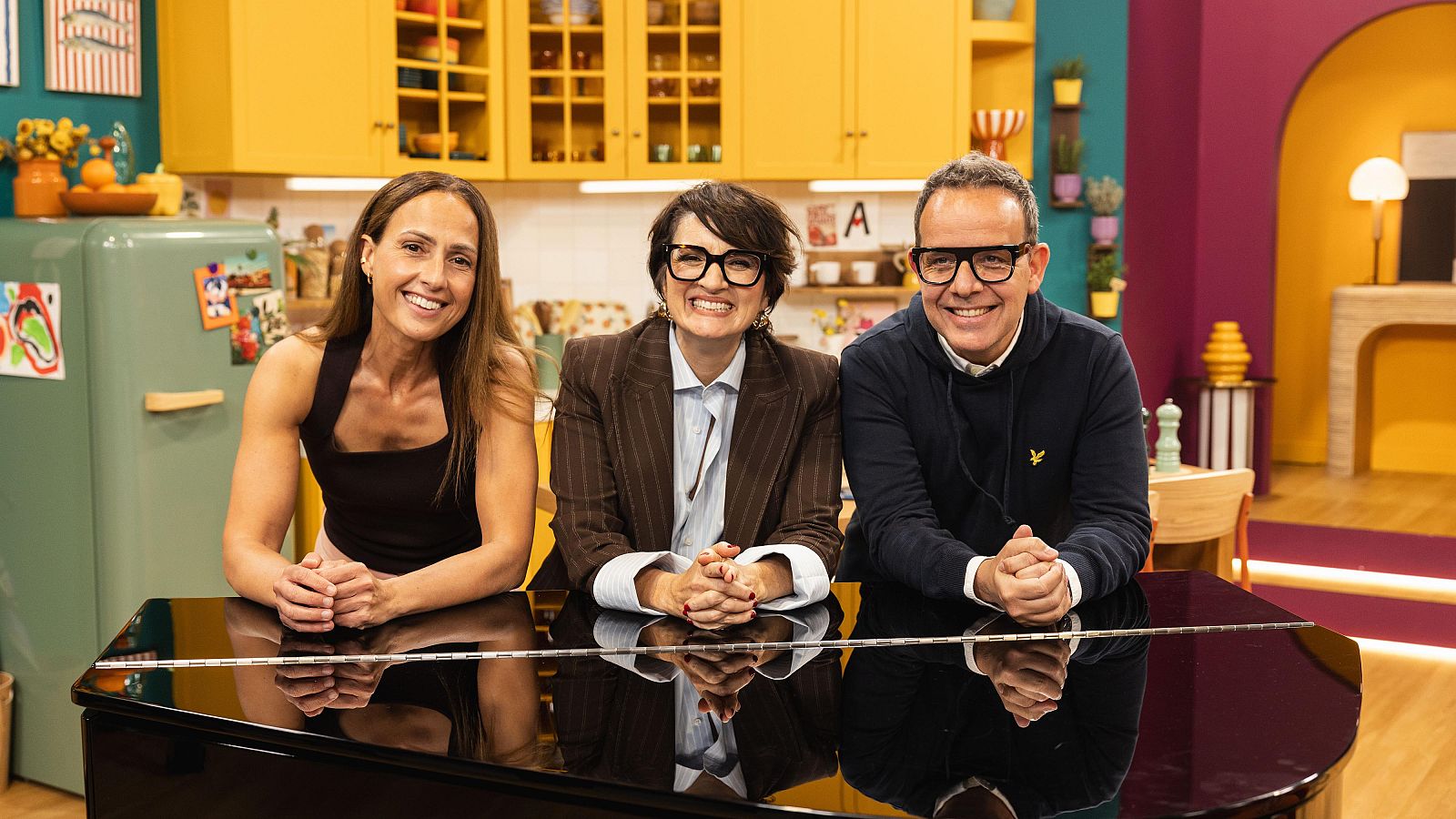 Tres persones somriuen i posen davant d'un piano negre. Una dona amb top negre, una altra amb ulleres i camisa blava, i un home amb ulleres i dessuadora blava, amb les mans entrellaçades. El fons és una cuina.