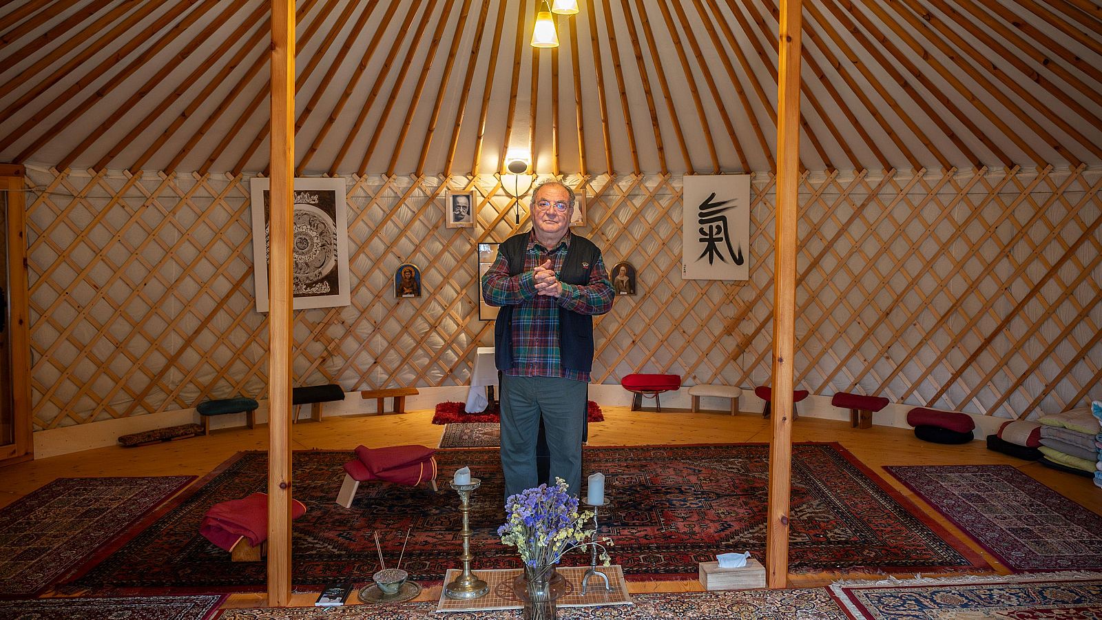 Josep Maria Fericgla, vestit amb camisa de quadres i armilla fosca, es troba a l'interior d'una estructura circular, possiblement un yurt, amb decoració oriental i símbols a les parets. Hi ha un ambient tranquil i espiritual.