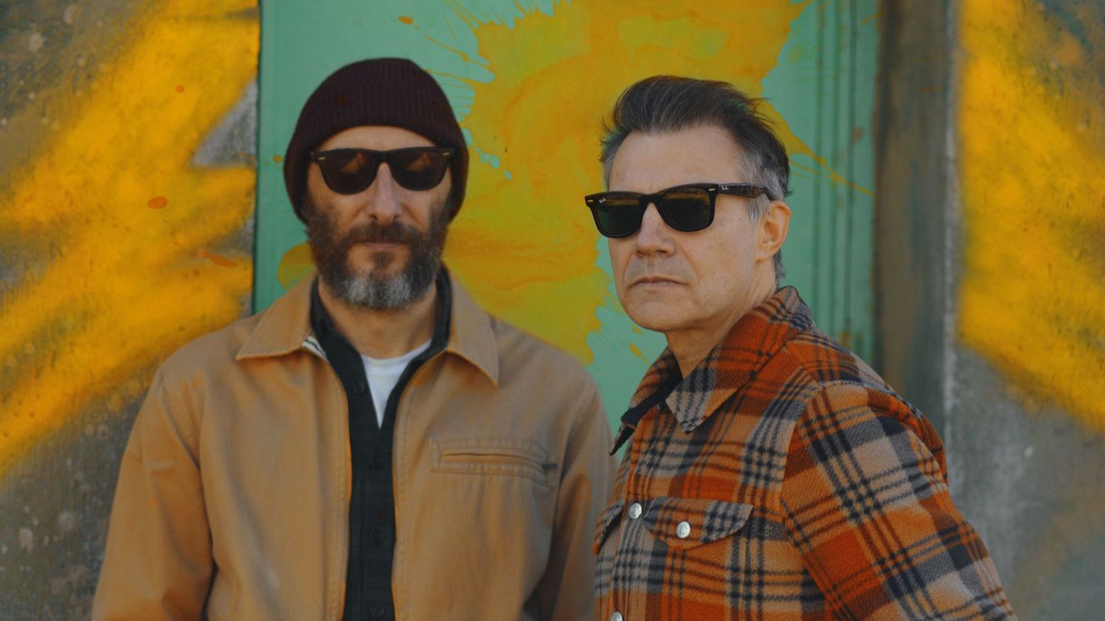 Dos hombres con gafas de sol posan delante de una pared con grafitis. Uno viste gorro y chaqueta clara, el otro una camisa de cuadros. En la parte superior se lee una inscripción relacionada con un evento.