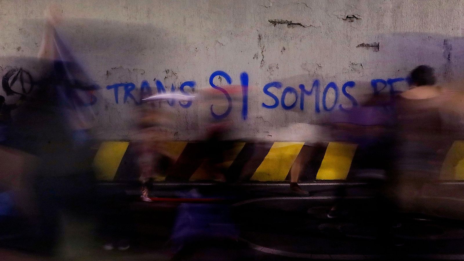 Imagen borrosa de gente caminando delante de un grafiti en el que se lee "Trans SI"