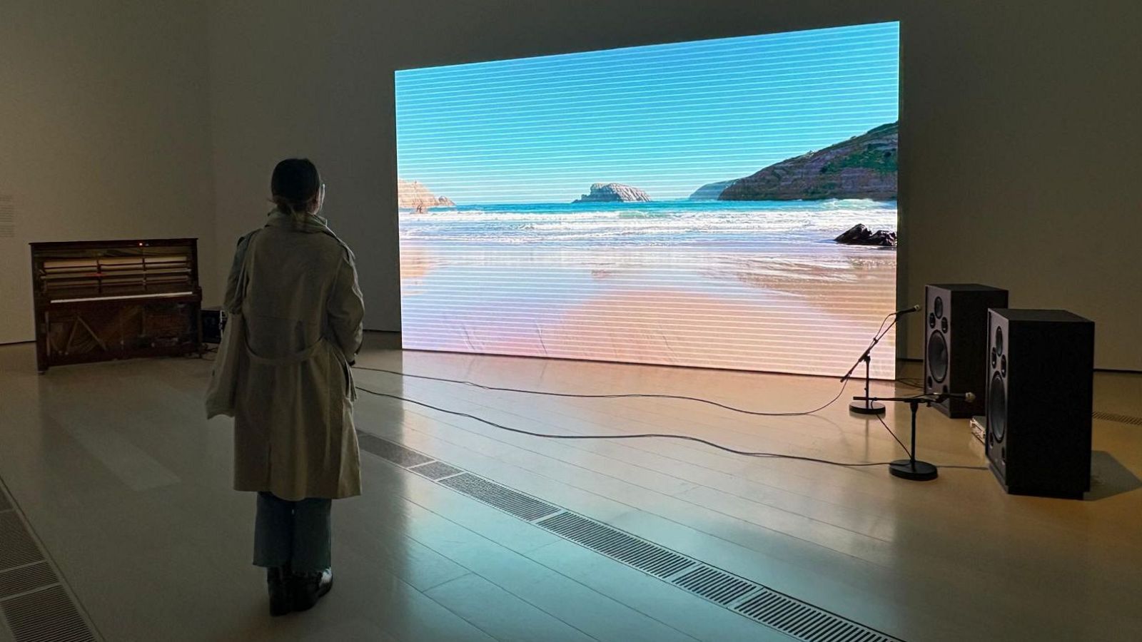 En una sala de exposiciones, una persona observa una proyección de una playa en una pantalla grande. A su lado, se encuentra un piano vertical y equipos de sonido, incluyendo altavoces y micrófonos.