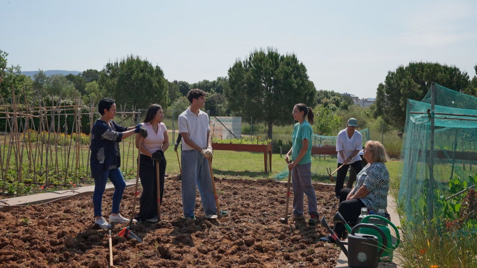 En un huerto, varias personas, vestidas con ropa informal, trabajan y conversan cerca de un terreno arado. Se observan herramientas de jardinería y vegetación en el entorno.