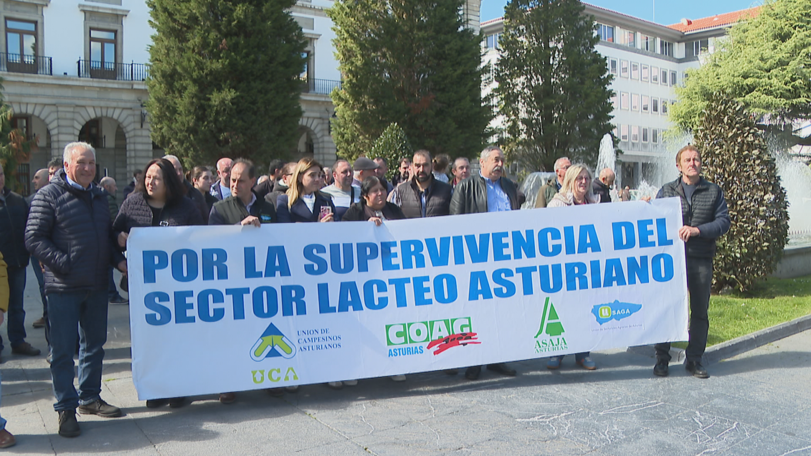 En una plaza, un grupo de personas protesta portando una pancarta con el mensaje "POR LA SUPERVIVENCIA DEL SECTOR LÁCTEO ASTURIANO", con logos de organizaciones agrarias. Se encuentran en un entorno urbano con edificios y una fuente.