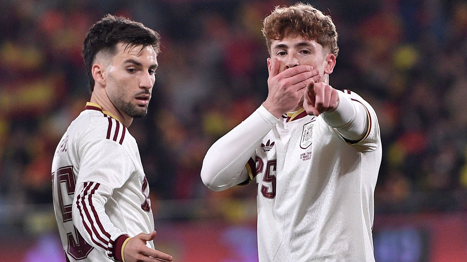 Dos jugadores de fútbol, Víctor Muñoz y Álex Baena, celebran un gol vistiendo la camiseta de la selección española, con Baena tapándose la boca y señalando con el dedo.