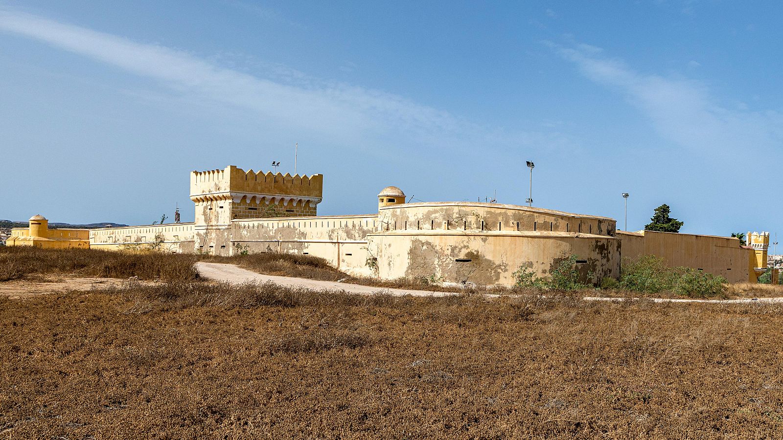 Vista panorámica de un complejo arquitectónico fortificado de tonos ocres y amarillentos, con terreno árido en primer plano y detalles como torre, muros con aberturas y garita de vigilancia.