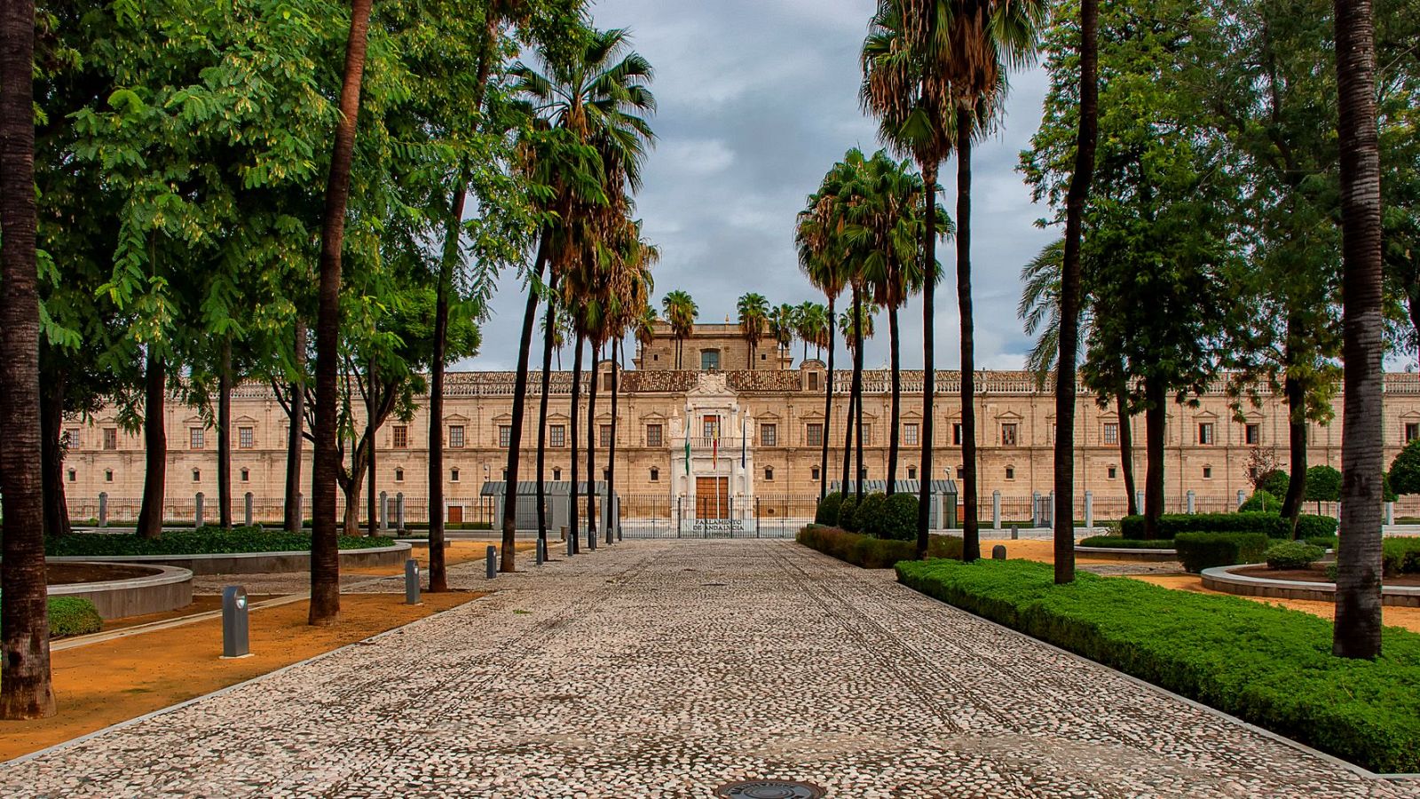 Un edificio de estilo clásico, con una entrada principal y banderas, se alza frente a un camino pavimentado. La escena se completa con palmeras y áreas ajardinadas, bajo un cielo nublado.
