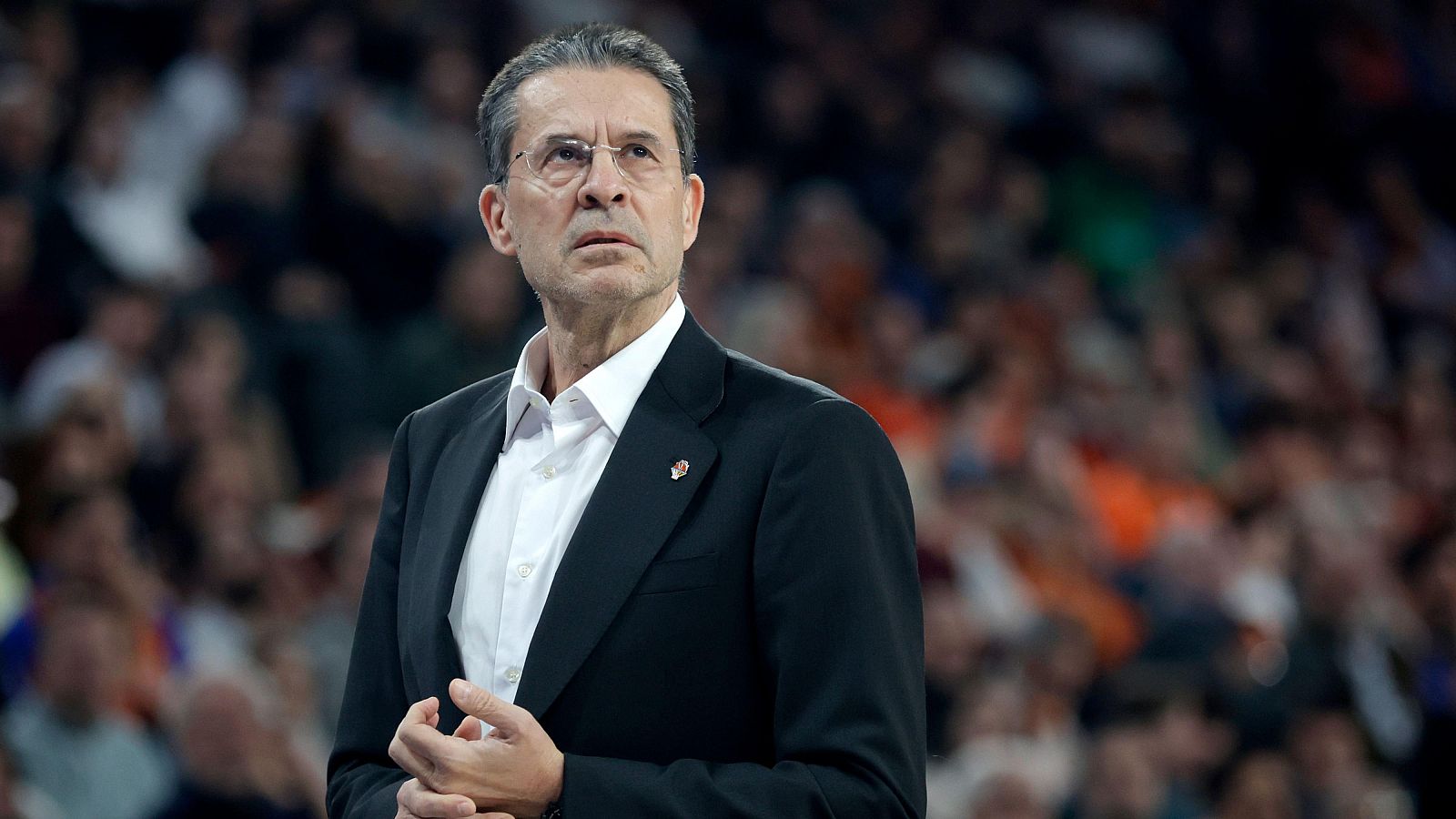 Pedro Martínez, entrenador del equipo de baloncesto, aparece serio y concentrado, vestido con camisa blanca y traje oscuro, con un pin en la solapa, mientras una multitud observa el partido.