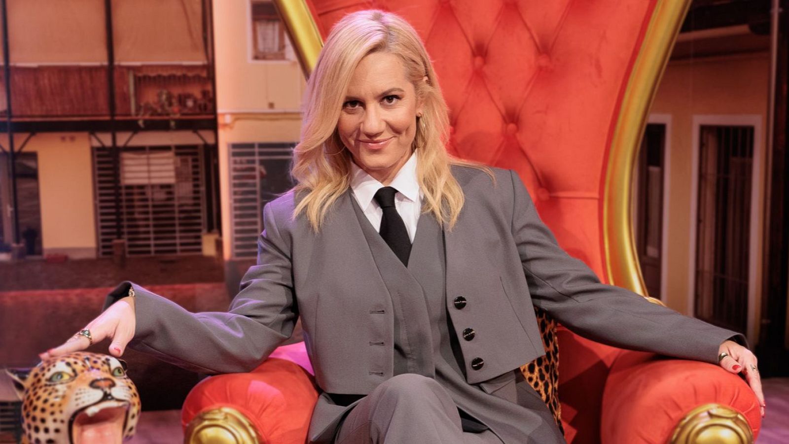 Henar Álvarez, presentadora de televisión, sonríe sentada en un sillón rojo. Viste un traje formal con corbata, y su mano descansa sobre una figura decorativa. El fondo muestra una escenografía que simula un edificio.