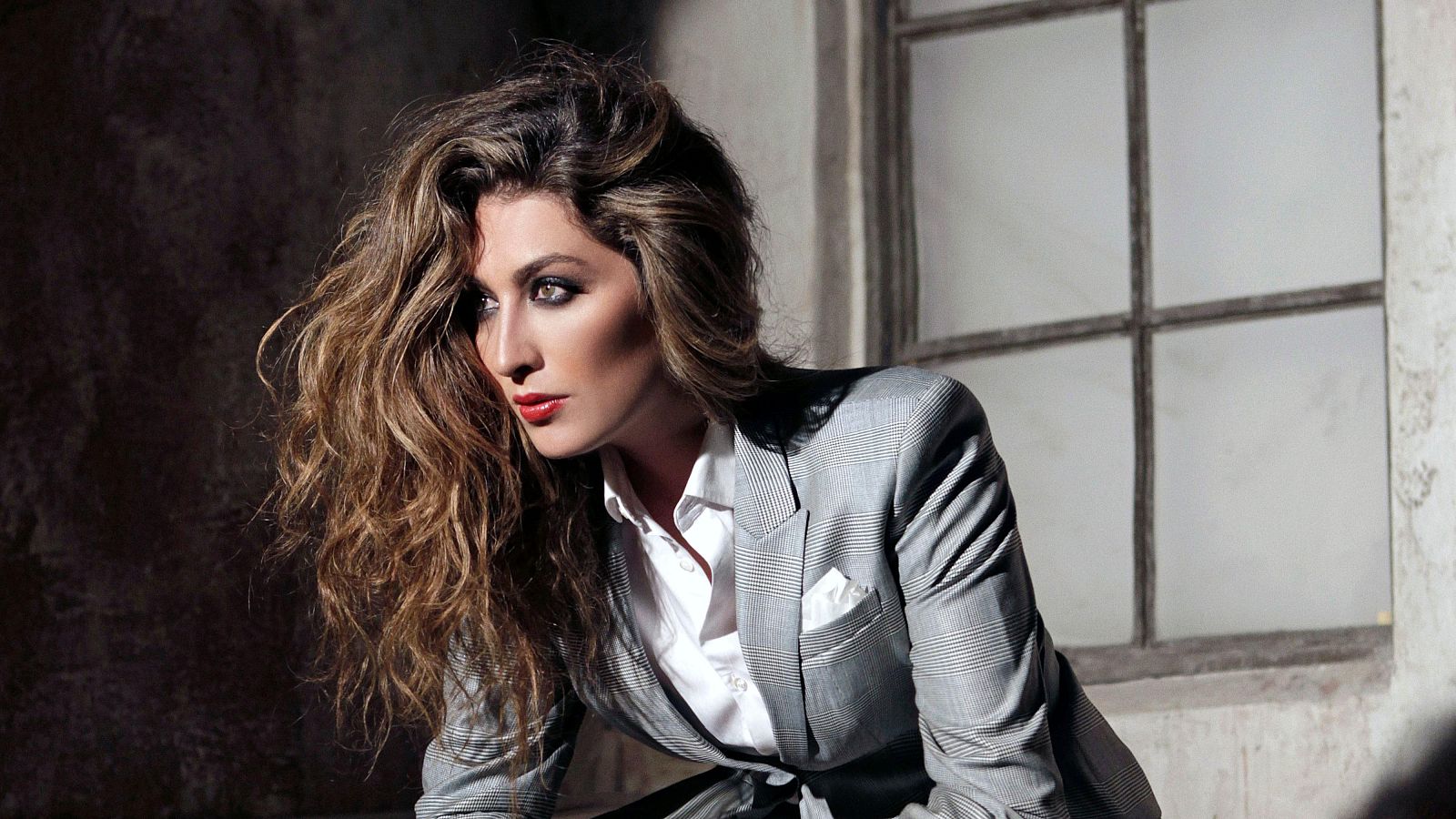 Estrella Morente posa con un traje gris a rayas y camisa blanca, luciendo su cabello castaño y ondulado. Destacan sus labios rojos y el maquillaje, con una ventana como fondo.