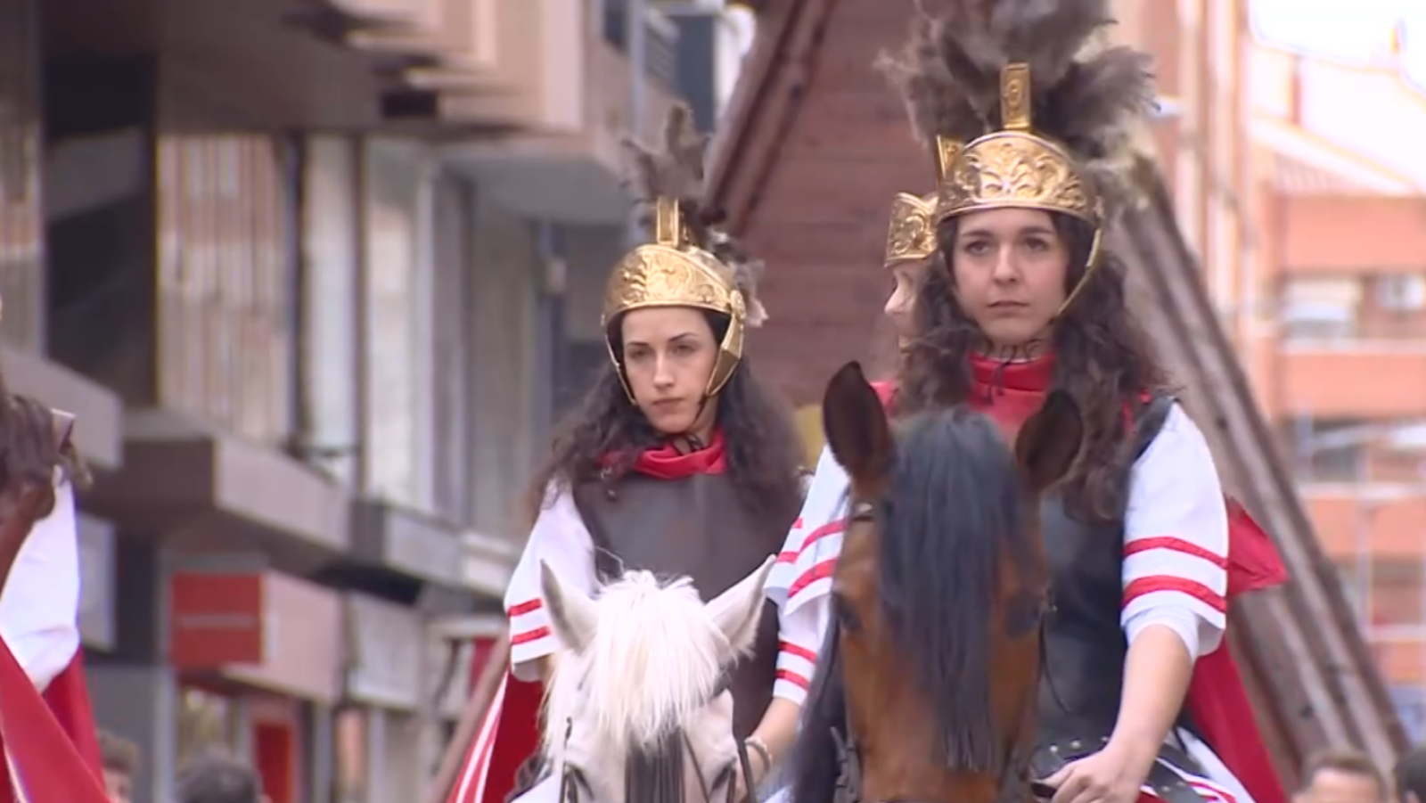 La imagen presenta a dos mujeres montadas a caballo, vestidas con atuendos que evocan la antigua Roma, incluyendo cascos dorados con plumas, capas rojas y armaduras.