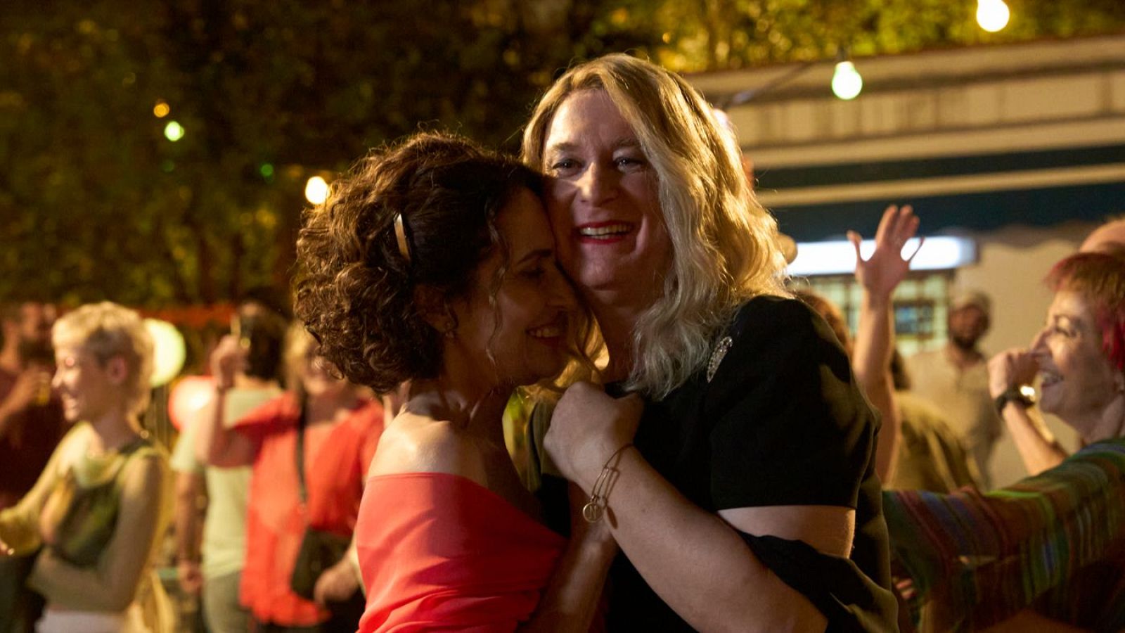 Dos personas se abrazan en una escena festiva nocturna. Un hombre con camisa negra y cabello rubio sonríe, mientras una mujer con vestido rojo y cabello rizado disfruta del momento, rodeados de luces y otras personas.