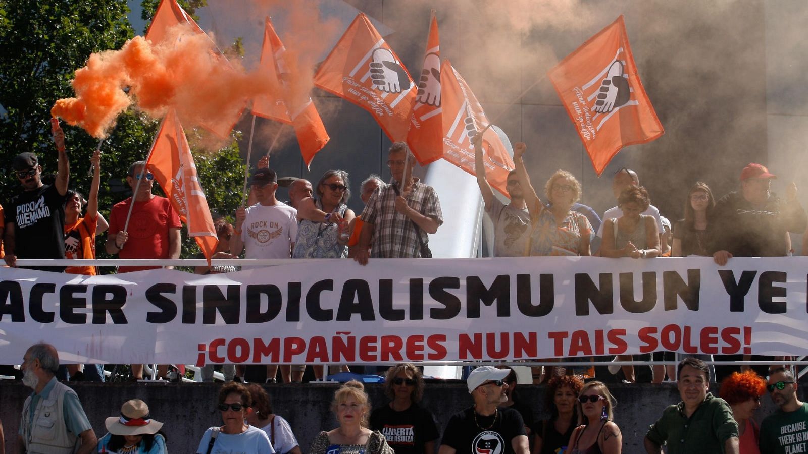 Una multitud se manifiesta tras una pancarta con un mensaje. Se observan banderas naranjas y humo, mientras algunas personas visten camisetas con inscripciones.