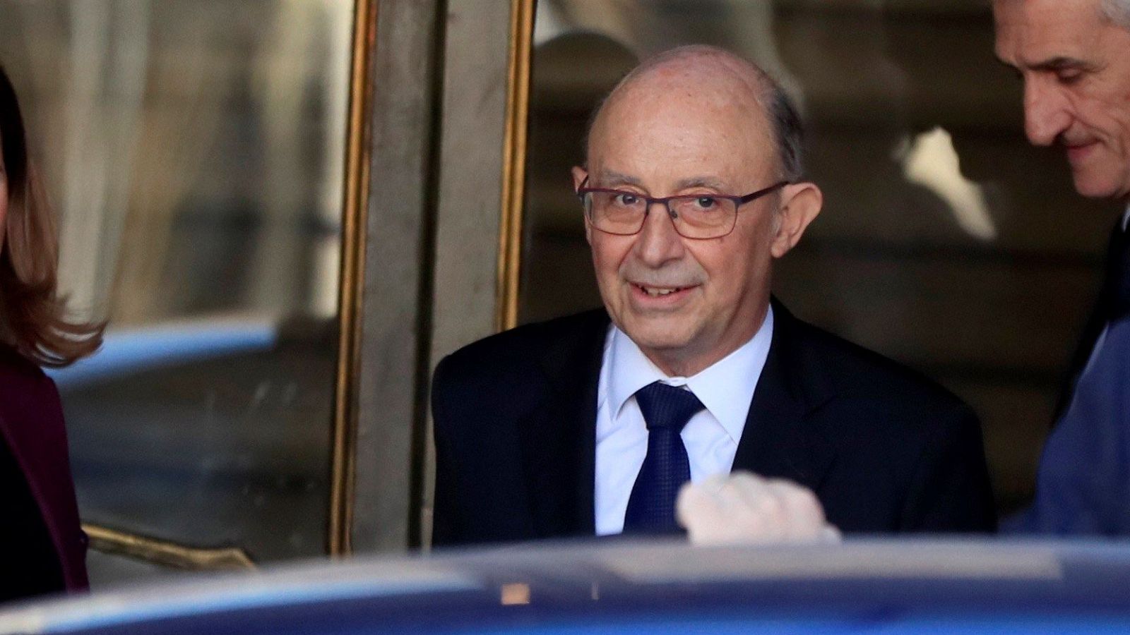 Imagen de Cristóbal Montoro, exministro, con traje oscuro y corbata azul, sonriendo ligeramente.  A su lado, partes visibles de otras dos personas. Fondo con puerta y coche.