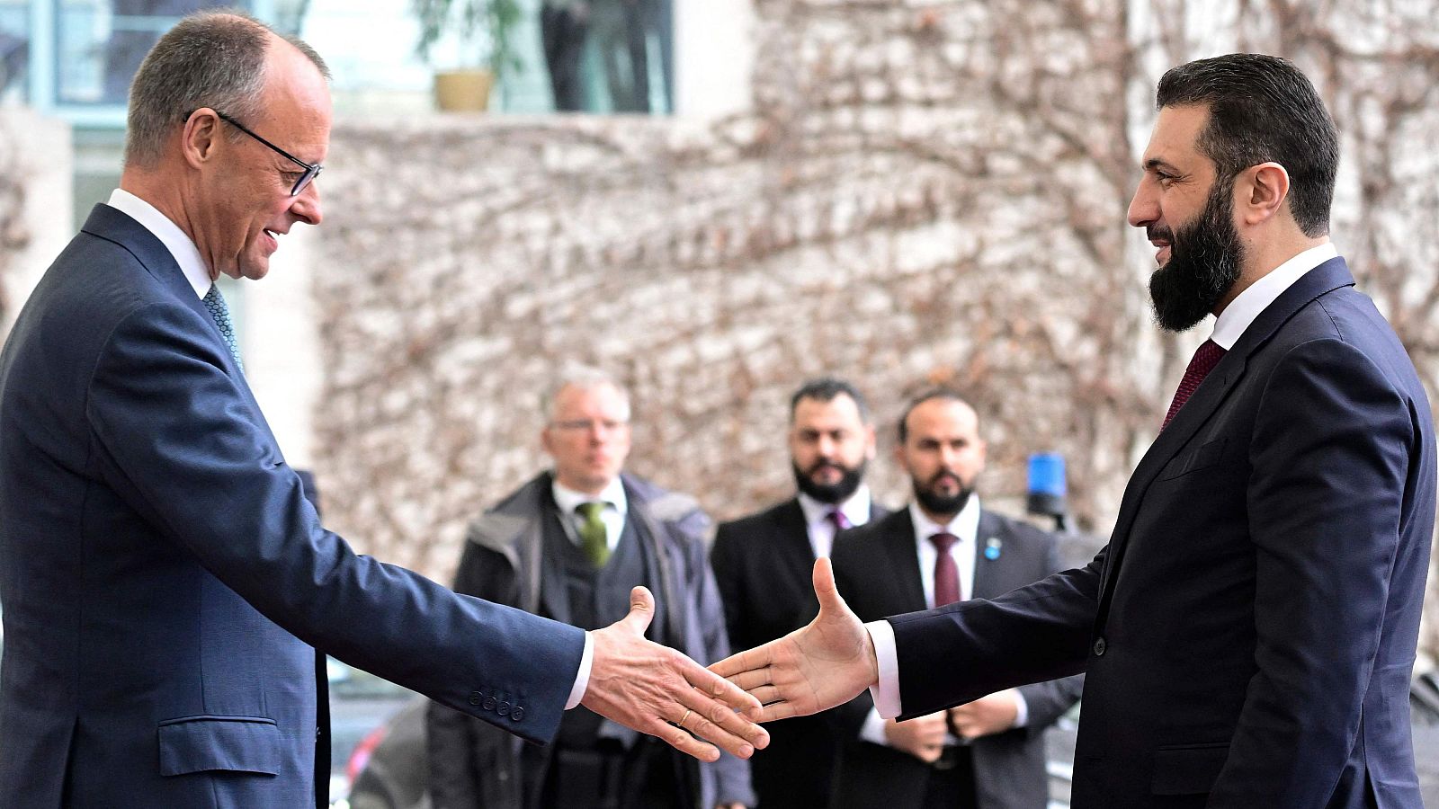 Friedrich Merz y Ahmed al-Sharaa se dan la mano frente a un edificio, ambos vestidos con trajes oscuros. Se observa a otras personas en el fondo, también con vestimenta formal.
