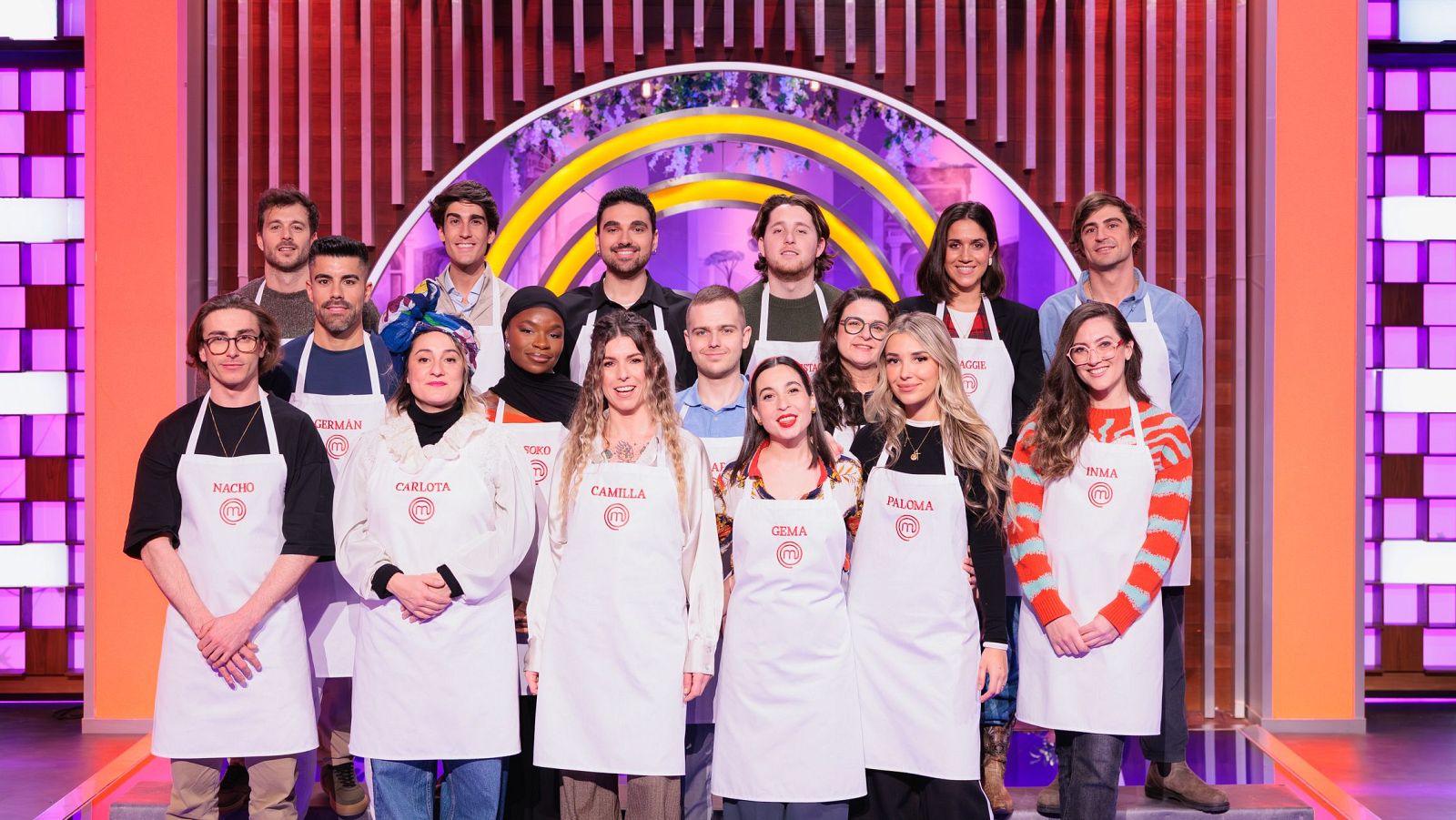 Un grupo de aspirantes a MasterChef, con delantales blancos, posan para una foto. Se pueden leer algunos nombres en los delantales, mientras que el fondo muestra una estructura decorativa con luces.