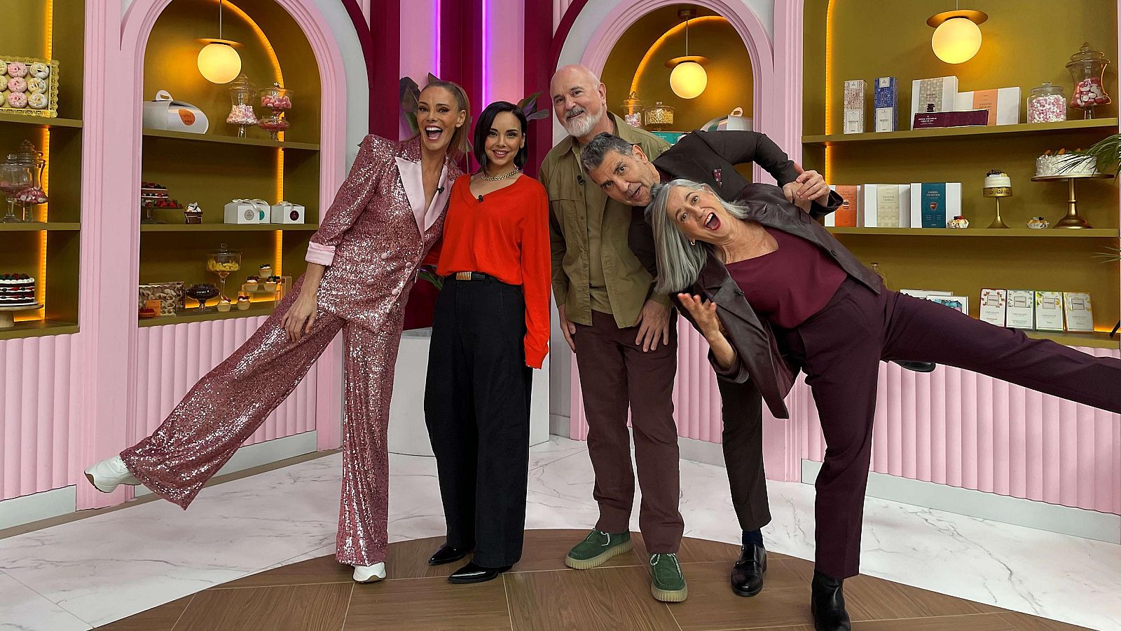 Cinco personas sonríen en un plató de televisión con decoración de pastelería. Una mujer con un traje rosa brillante y coleta alta destaca en la imagen, rodeada de otras dos mujeres y dos hombres, todos posando frente a estanterías repletas de dulces.