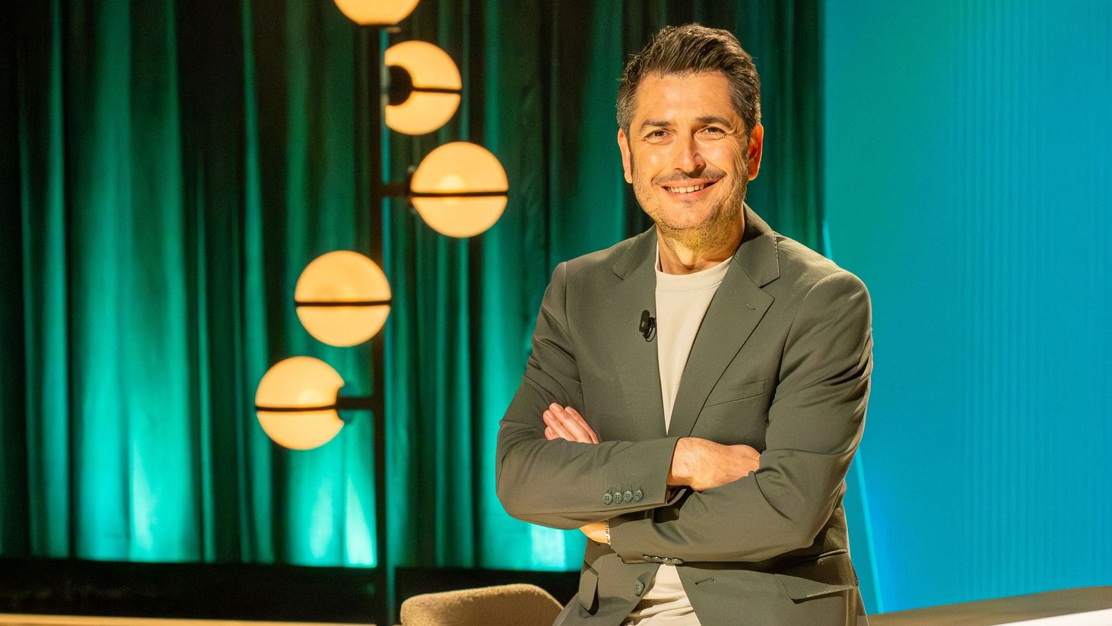 Carlos del Amor, con el pelo canoso y una sonrisa, posa con los brazos cruzados, vestido con una chaqueta gris y camiseta blanca, frente a un fondo con cortinas, una lámpara y una pared azul.