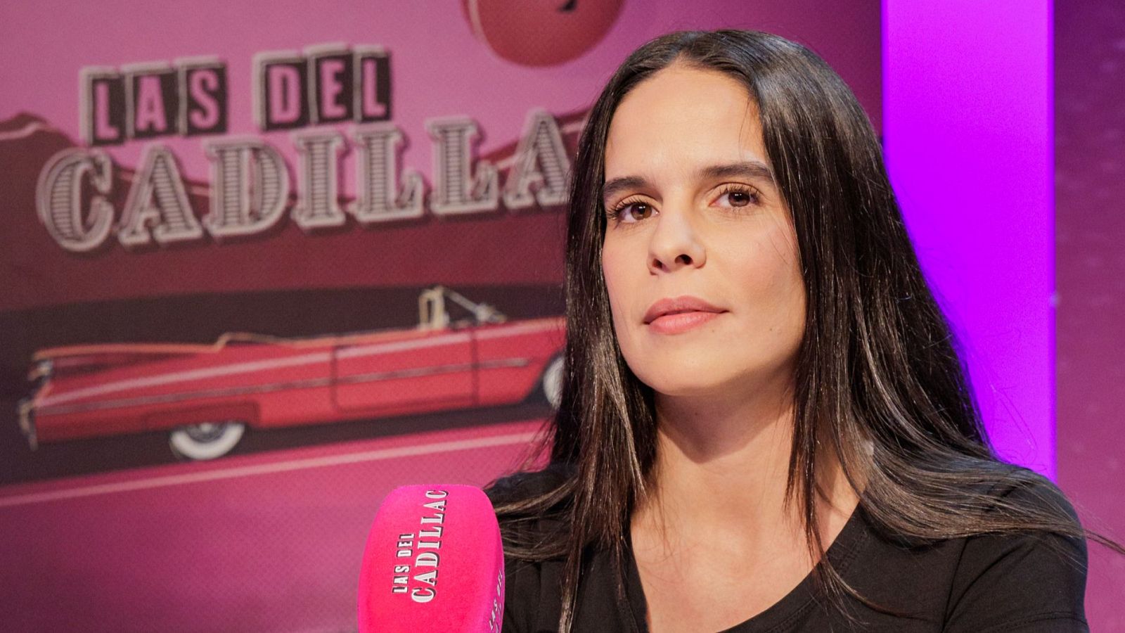 Ángeles Toledano, con pelo oscuro y canas, se encuentra frente a un fondo rosa con el logo de un programa que incluye un coche rojo. Un micrófono rosa con el nombre del programa está presente, posiblemente en un estudio.