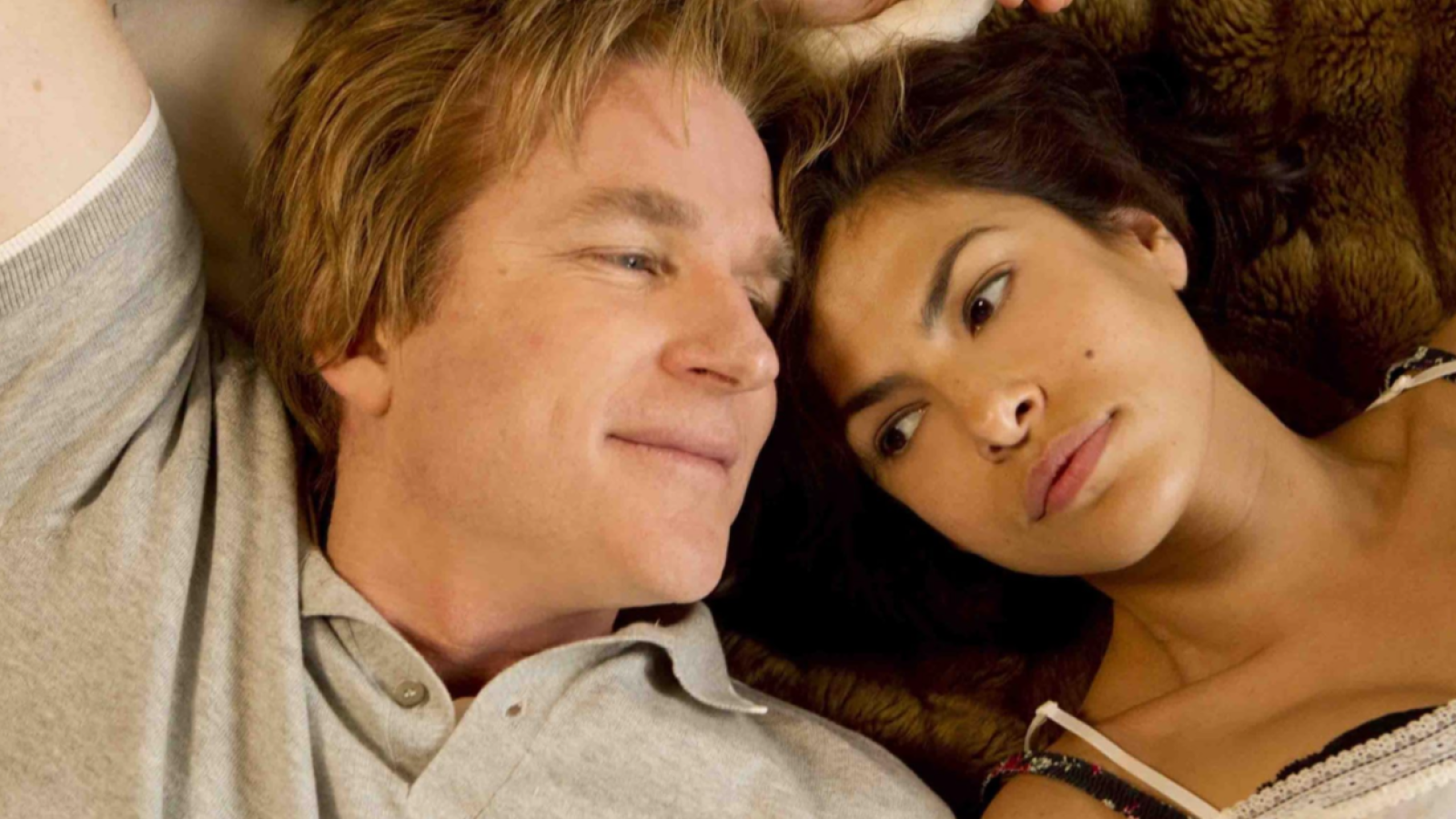 Matthew Modine y Eva Mendes, recostados juntos, comparten una mirada. Él, con camisa gris, sonríe levemente; ella, con detalles florales en su vestimenta, parece pensativa.