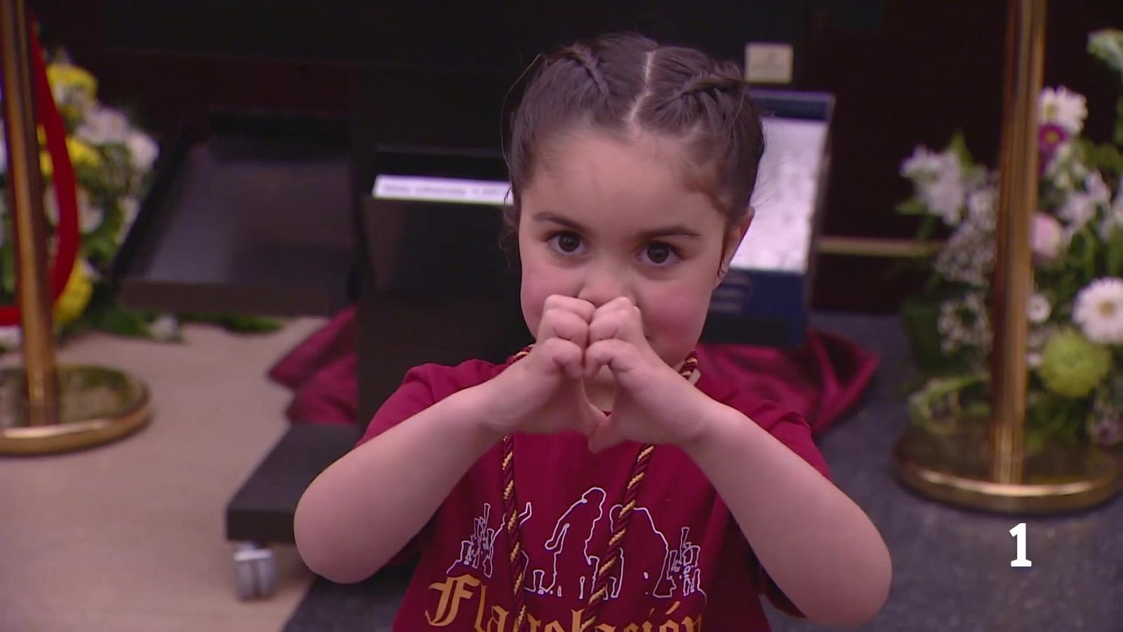 Una joven con trenzas, camiseta roja y un collar con los colores de una bandera regional, hace un gesto de corazón con sus manos mientras mira a la cámara, con un atril y flores de fondo.