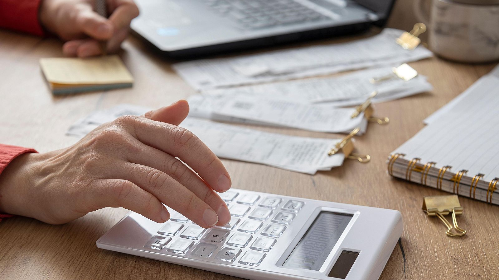 Una mano opera una calculadora blanca, rodeada de recibos, facturas y elementos de oficina. Al fondo, se distingue un ordenador portátil abierto, una libreta y notas adhesivas, indicando un entorno de trabajo y cálculo.