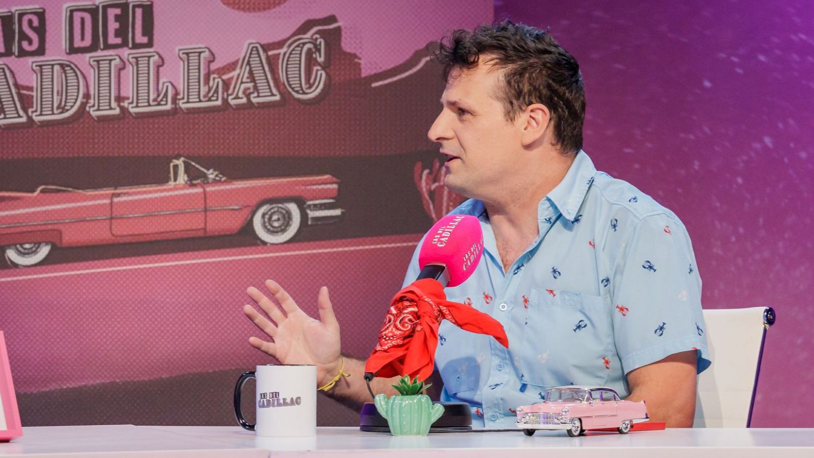 Ángel Carmona, en un estudio, gesticula mientras está sentado frente a una mesa con un micrófono, una taza y una maqueta de coche. El fondo muestra el nombre de un programa y la imagen de un coche clásico.